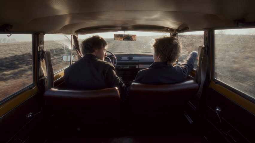 Young couple traveling in vintage car through countryside, chatting and laughing during romantic road trip. View from behind