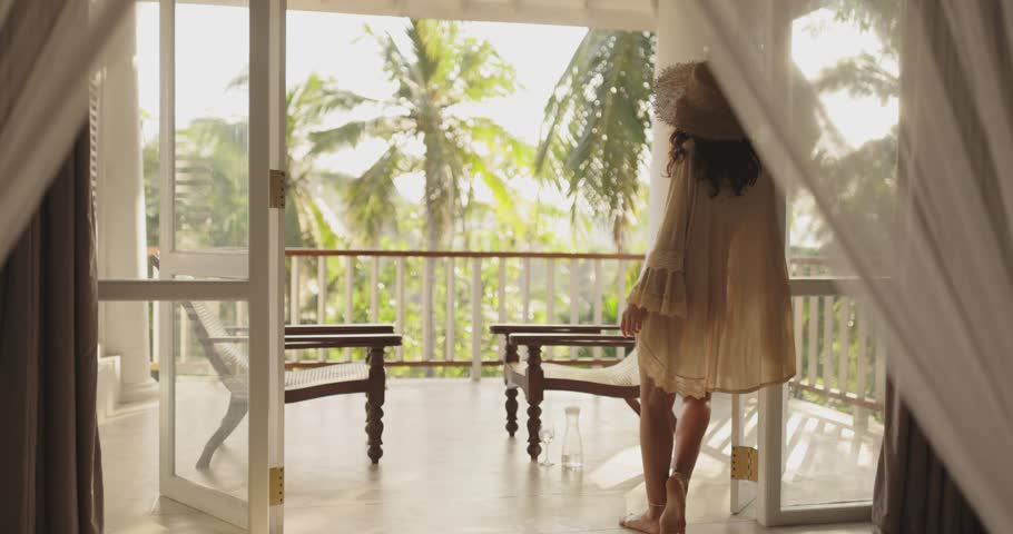 A woman stands gracefully on a tropical balcony, framed by sheer curtains, enjoying the lush greenery. Perfect for luxury lifestyle, travel promotions, and exotic vacation concepts.