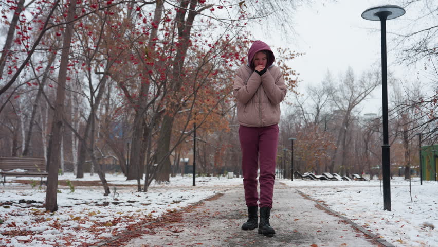 Woman in winter outfit strolling alone, adjusting her hood and bringing her hands close to her mouth to warm up, snow-covered ground, leafless trees, and scattered autumn leaves