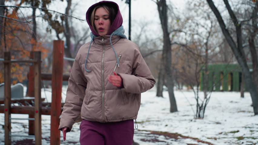 Young woman in winter outfit walking briskly through snowy park in urban setting, warming up in cold weather, trees with no leaves in background