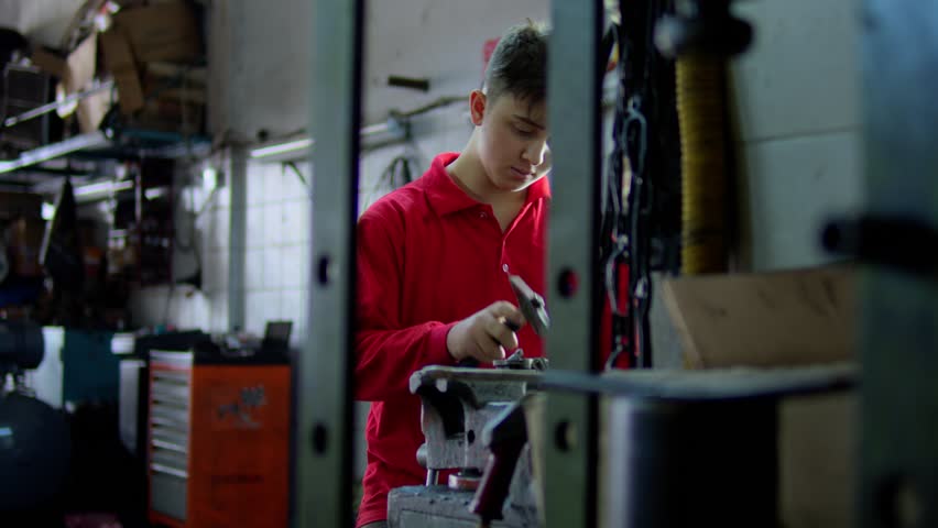 Young mechanic working in an auto repair shop, using a tool on a metal part. Focused apprentice learning mechanical skills in a workshop environment with industrial equipment.