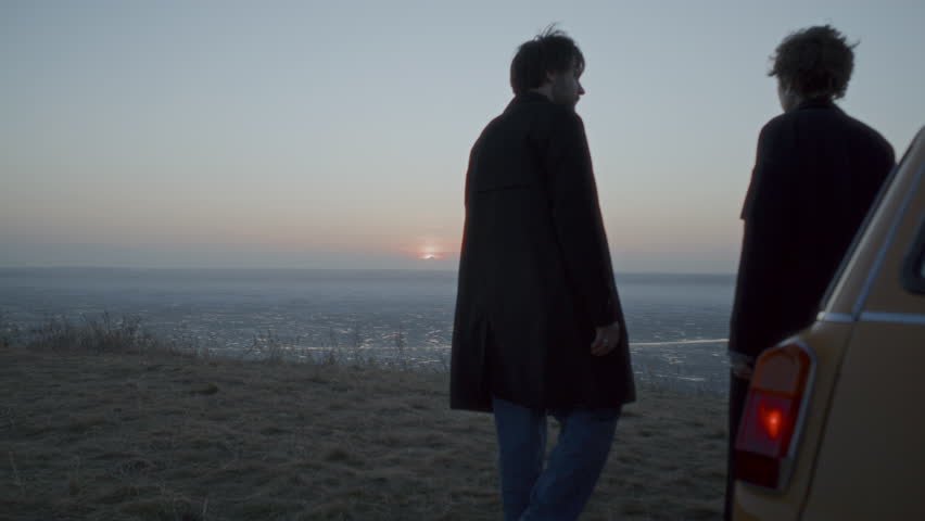 Young couple in love walking away from parked car, holding hands, looking at scenic sunset over frozen lake during stop on romantic road trip. Rear view