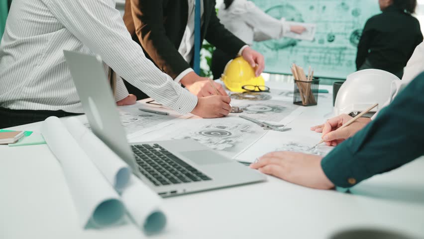 A diverse team engaged in a collaborative engineering project, focusing on detailed drawings and tools while ensuring safety with helmets in a modern workspace. SACTR