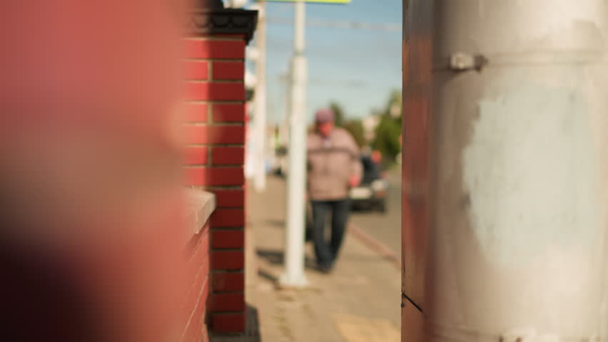 Blurry man walking along city sidewalk beside red brick wall under bright sunlight with soft focus vintage film look and shallow depth of field creating mysterious urban atmosphere