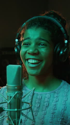 Vertical portrait of joyful African American teenage musician looking at camera while recording song with headphones in front of studio microphone