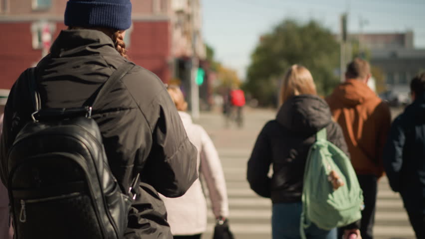 Group of people walking on urban street crossing on sunny day. Pedestrians wearing jackets, backpacks, heading towards city. Focus on individuals walking and urban surroundings