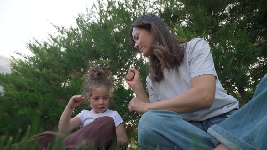 Happy mother and child enjoying time together outdoors, sitting on grass and playing rock paper scissors hand game