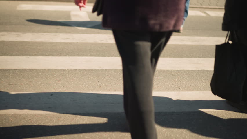 Close-up shot of lower bodies of people walking across road on busy day, with shadows cast on pavement, showcasing shoes and movement in urban setting, highlighting pedestrian traffic