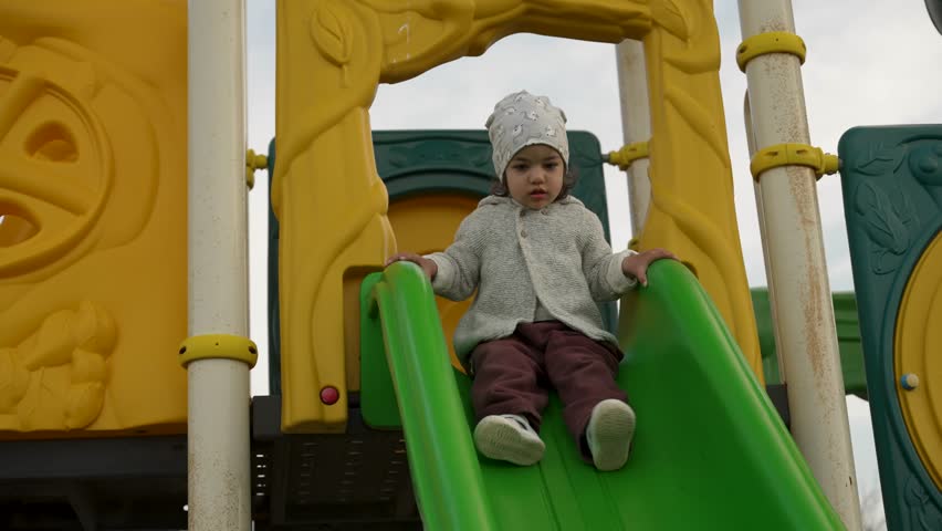 Toddler Girl Sitting at the Top of a Playground Slide. A cute baby wearing a hat and sweater sits at the top of a slide on a colorful outdoor playground, looking down