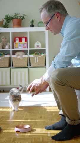 Middle-aged man feeding a fluffy exotic shorthair cat at home sitting in a cozy sunlit living room, gently petting it while the cat eats from a pink bowl on the floor.