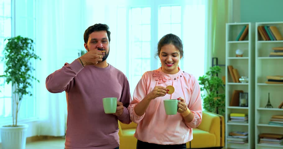 Indian Asian young couple happily enjoying hot tea coffee with biscuits, standing together in cozy living room, smiling bonding over morning beverage, sharing warmth, love, and relaxation at home
