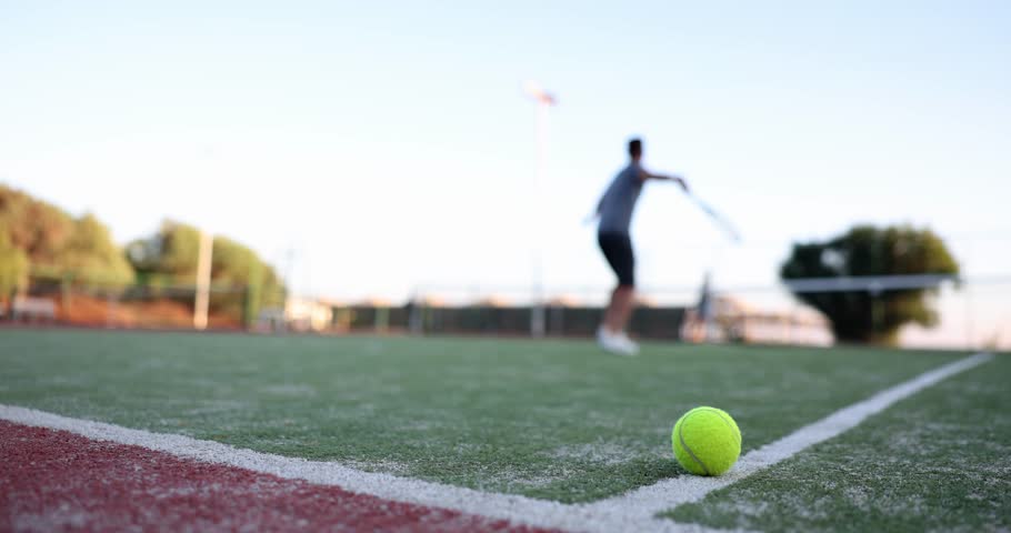 Bright yellow ball against male person playing tennis on grass playground. Man jumps to hit ball on outdoor tennis court focusing on active sport