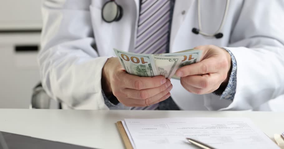 Doctor in coat and stethoscope around neck counts dollar bills sitting at desk. Medical documents suggest healthcare costs and financial aspects