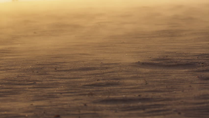 Golden sunset casting warm light over arid desert landscape, wind sweeping fine sand grains and creating dynamic, flowing patterns across desolate terrain