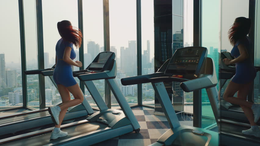 Young woman in sportswear runs on treadmill in gym. Strong sportswoman exercises on modern trainer in inspiring workout environment. Lady enjoys sports activity in urban area