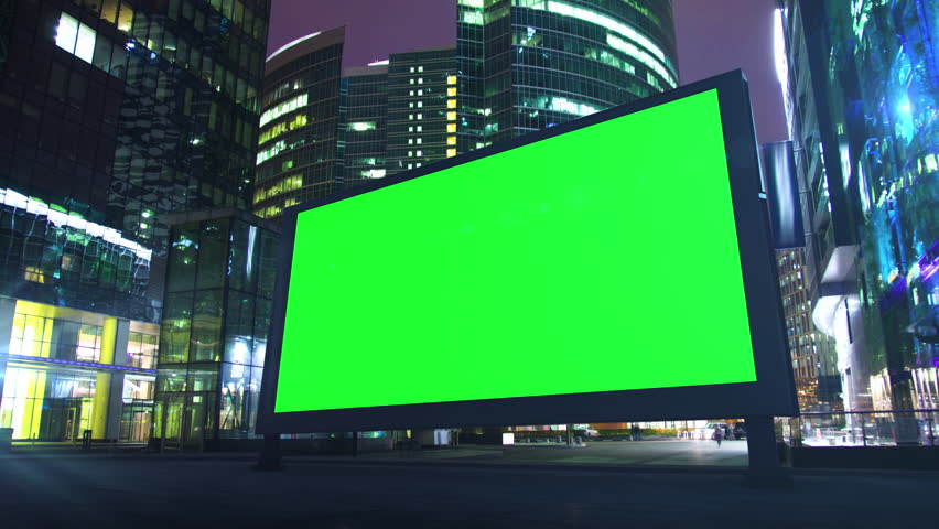 Blank green screen billboard stands between modern corporate buildings with business people walking by at night