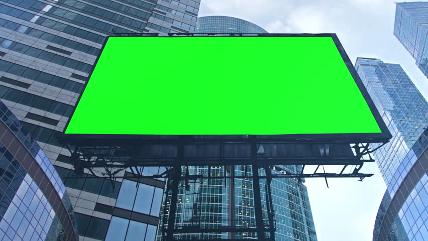 Large blank green screen billboard is hanging between modern glass skyscrapers