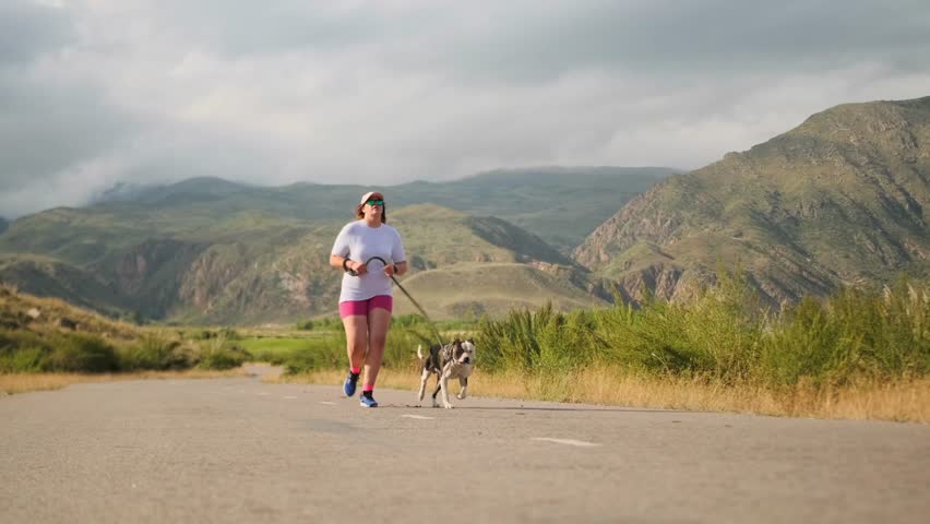 An overweight teenage girl in sportswear runs with a pit bull along a mountain road under a cloudy sky. The dog runs happily beside her, panting and matching her pace, adding energy to their train.