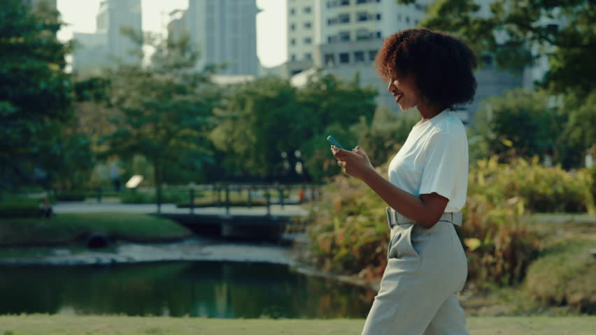 Happy African American lady with smartphone walks past park artificial pond. Pretty young woman scrolls social media content via mobile phone in urban environment side view