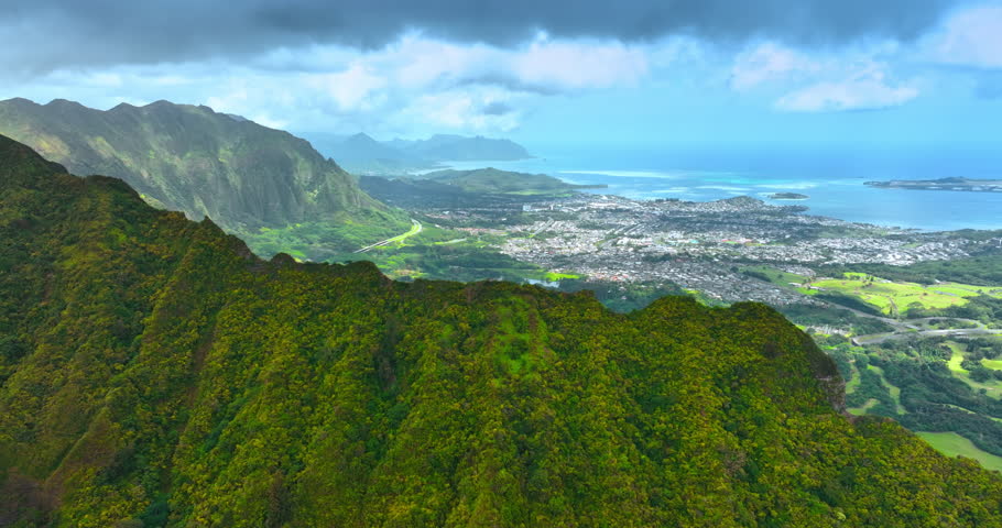 Rising over the stunning verdant mountain range. Revealing view of Honolulu cityscape under the dramatic cloudscape. Azure waterscape at backdrop.