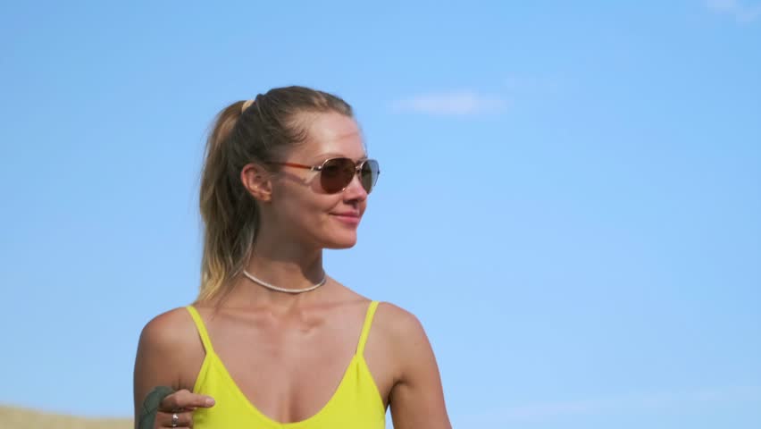A fit young woman in a yellow top, white bike shorts, and sunglasses walks a pit bull along a desolate mountain road. Dry hills and sparse bushes stretch under bright summer sunlight.