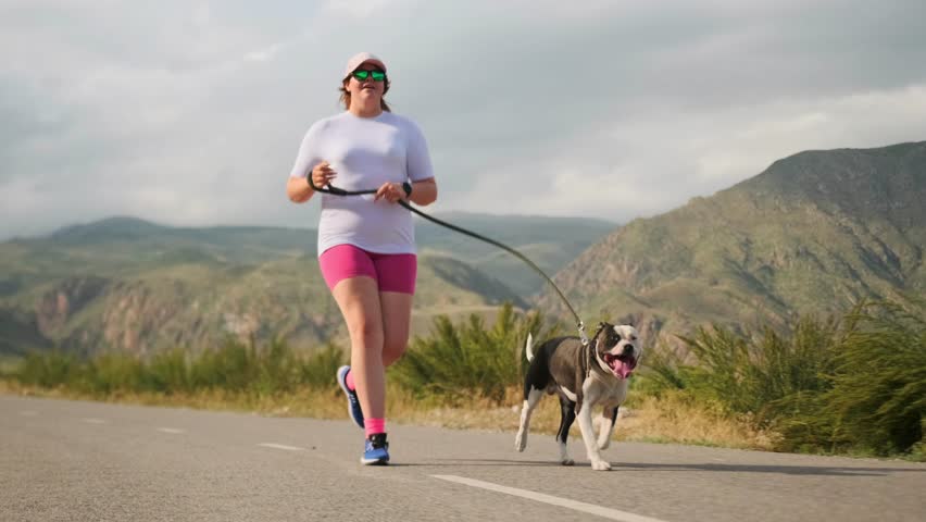 Long shot. An overweight teenage girl in sportswear runs with a pit bull along a mountain road amid green hills. The dog stays beside her, panting, as she pushes to lose weight and build endurance.