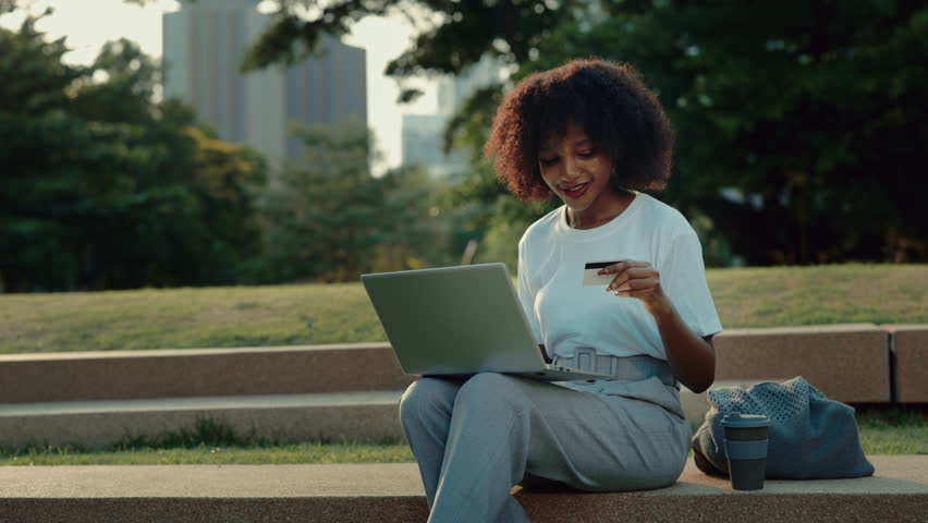 Portrait of African American woman paying online with credit card via laptop in park. Young lady uses safe money transfer on internet sitting outdoors. Digital banking