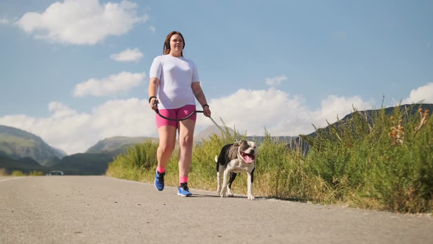 Low angle. An overweight teenage girl in sportswear walks with a pit bull along a brush lined road, mountains ahead. Under the midday sun, they walk as the dog pants, trying to cool off in the heat.