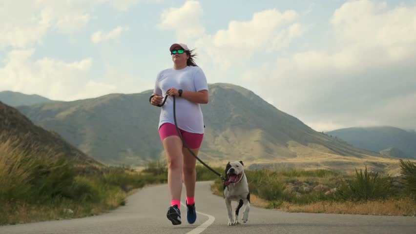 Long shot. A young woman in sportswear runs with a pit bull along a mountain road past green hills. Her dog stays close, helping her push through fatigue and stay focused on her weight loss goal.