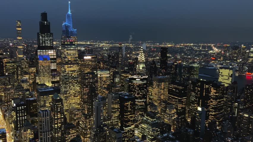 Aerial pan reveals illuminated Midtown Manhattan skyscrapers at night. Includes Bank of America Tower, Chrysler Building. Urban energy, NYC nightlife, business, establishing shot, USA