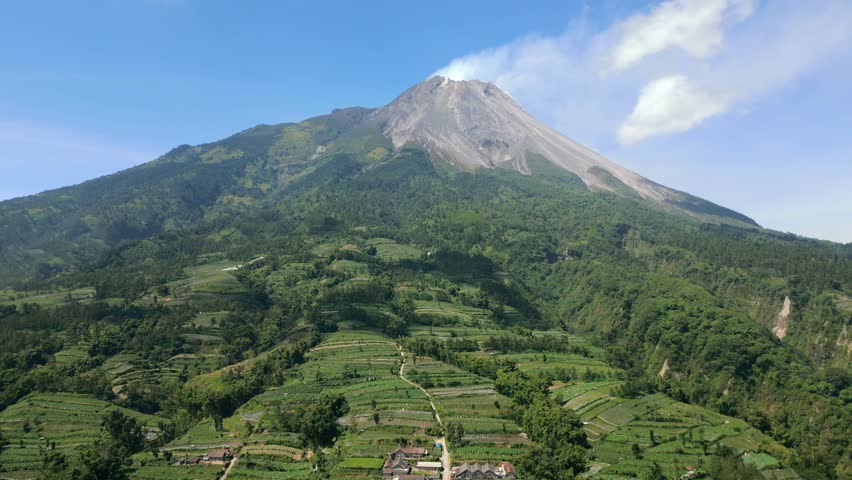 Aerial view of Mount Merapi volcano surrounded by lush green farmland and rural landscape in Java, Indonesia. Scenic volcanic terrain and cultivated hills under clear blue sky.