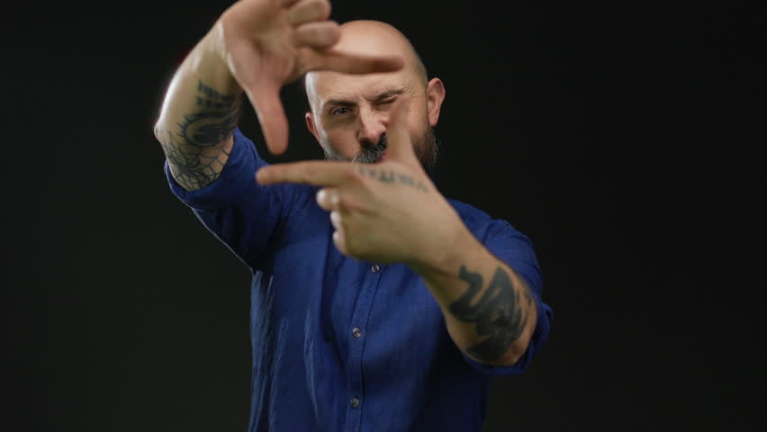 Man with tattoos making frame gesture with hands in front of face against black background suggests creativity and focus