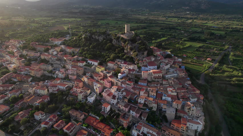 Posada, Sardinia, at sunset, featuring Castello della Fava atop a rocky hill, with its medieval village spreads below, surrounded by lush fields, olive groves, slow motion drone pulling out