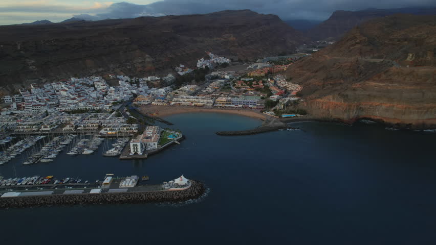 Drone view of Puerto de Mogán in Gran Canaria. The marina, golden beach, and whitewashed buildings sit beneath rugged hills, with calm waters and twilight sky enhancing the serene coastal atmosphere.