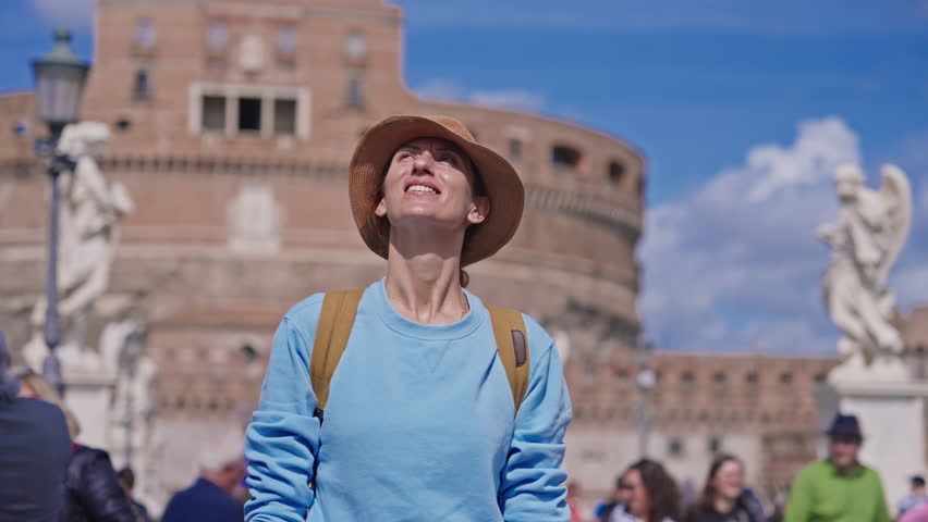 A girl tourist in a hat and with a backpack stands against the background of Saint Angel Castle on the bridge Ponte Sant Angelo over the Tiber river looking at the sights of Rome, Italy