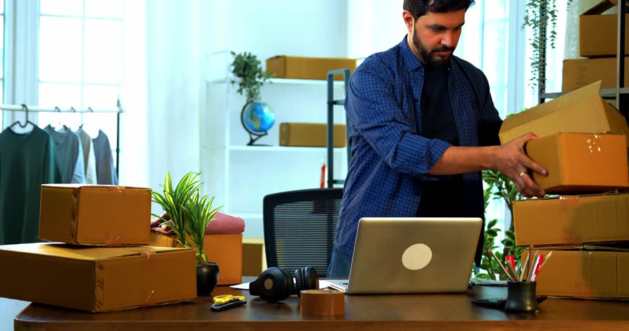 Indian Asian small businessman in store room office packing garments into cardboard carton box on desk for shipment, preparing online export dispatch order for delivery logistics