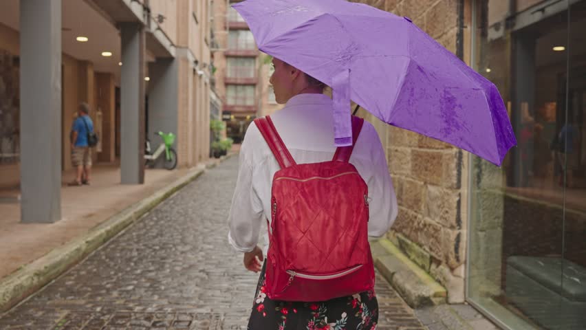 A young female tourist with a purple umbrella and red backpack walks along the cobblestone streets of Sydney's historic The Rocks district.