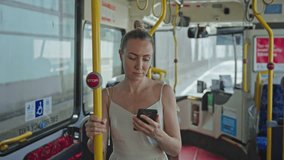 A woman in sunglasses and a light-colored dress is standing inside a public bus, holding a yellow handrail with one hand and a smartphone in the other. - Powered by Shutterstock - Get 15% off with code: PIKWIZARD15