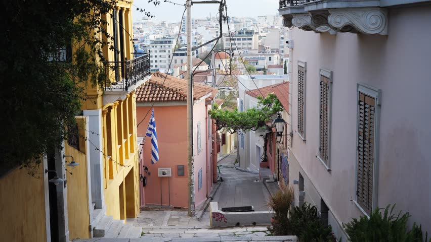 Narrow street in Athens, Greece, located in the historic old town tourist area. The street slopes downhill, with balconies, a hanging Greek flag, and a church visible in the background.