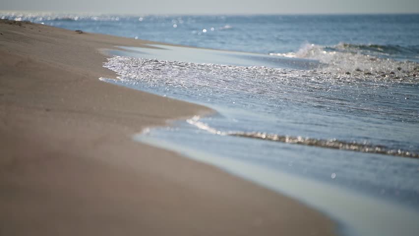 Calm ocean waves gently touch smooth sand at low tide. Sunlight highlights ripples and creates peaceful mood. Perspective shows shoreline extending into distance