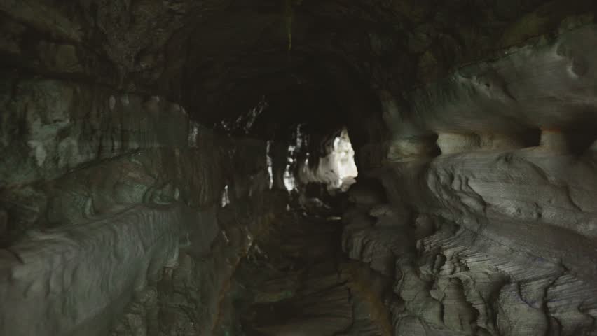 First-person view of narrow stone cave passage with visible light at the end. Mysterious underground tunnel with rough rocky walls and natural illumination.