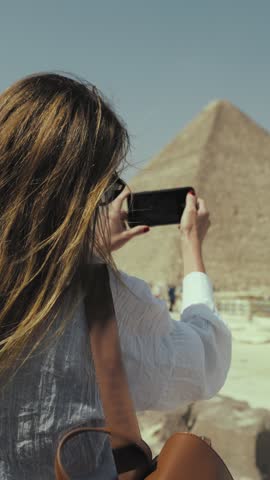 Happy woman tourist photographing pyramids in giza, mobile photo for memories, summer trip to Egypt. Excursion for exploring ancient history, culture and architecture, back view, female traveler