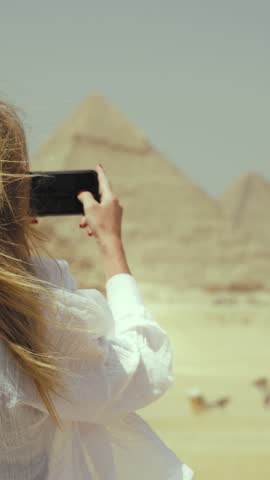 Carefree female tourist photographing beautiful majestic pyramids in Giza, Egypt. Popular tourist destination, attraction and landmark of ancient civilizations culture, back view of lady taking photo