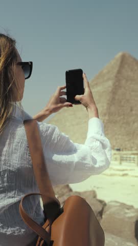 Tourist in Egypt, woman photographing famous pyramids by smartphone for memories, happy moments of summer vacation. Traveling to majestic landmarks in Giza, mobile photo for social networks, back view