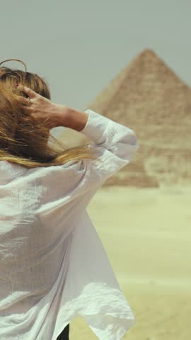 Blonde woman inspired by majestic pyramids in Giza stroking her swaying hair by wind, back view. Fleeting nature of human life and eternal pyramid, explore ancient culture and history of Egypt people