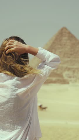 Blonde woman inspired by majestic pyramids in Giza stroking her swaying hair by wind, back view. Fleeting nature of human life and eternal pyramid, explore ancient culture and history of Egypt people
