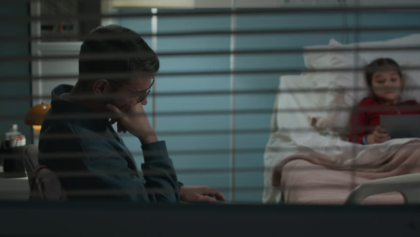 Handheld shot of tired father taking nap in chair while visiting sick daughter playing digital tablet in hospital ward