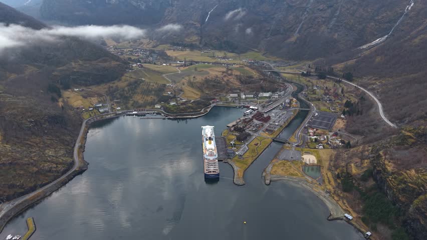 4K aerial drone view of Flam, Norway. A large cruise ship docks peacefully in the fjord, surrounded by majestic mountains, waterfalls, and the charming Nordic village landscape.