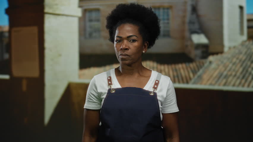 Woman wearing an apron indoors at an old university making a watching gesture with serious expression against a historic architecture backdrop.