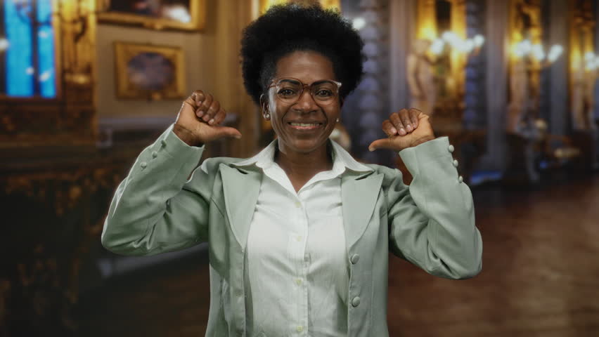Young african american woman in a museum pointing at herself with thumbs in an indoor setting, smiling confidently and surrounded by elegant artwork and lighting.